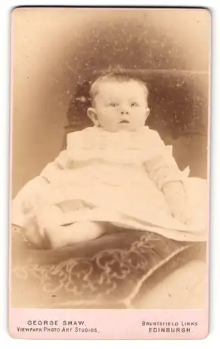 Fotografie George Shaw, Edinburgh, Portrait niedliches Baby im weissen Kleid auf Kissen sitzend