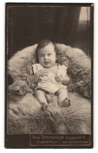 Fotografie Aug. Seringhaus, Elberfeld, Portrait dunkelhaariges Baby im weissen Hemdchen auf Fell liegend