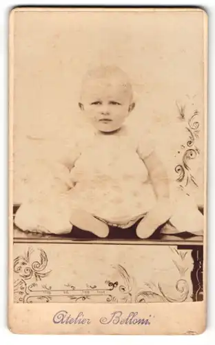 Fotografie Atelier Belloni, Ort unbekannt, Baby sitzt auf Kommode
