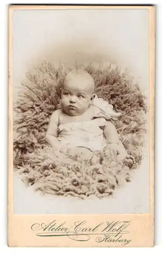 Fotografie Carl Wolf, Harburg a. E., Portrait niedliches Baby im Hemdchen auf Fell liegend