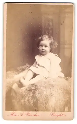 Fotografie Monsieur A. Boucher, Brighton, Baby im Kleidchen auf Pelzdecke sitzend