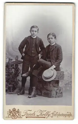 Fotografie Friedr. Kolby, Zwickau, Junge stehend im Anzug und Junge sitzend im Anzug mit Hut in der Hand