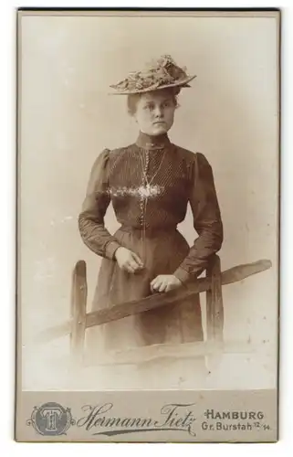 Fotografie Hermann Tietz, Hamburg, Portrait Dame in hübscher Kleidung mit Hut an Zaun gelehnt