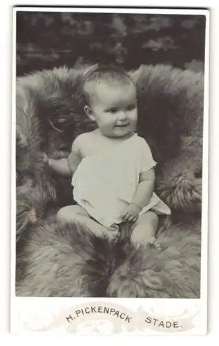 Fotografie H. Pickenpack, Stade, Baby im Kleidchen auf Pelz sitzend