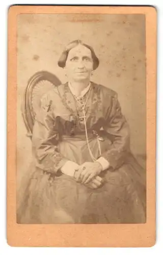 Fotografie Friedrich Baechler, Weissenfels, alte Dame im Kleid auf Stuhl sitzend mit Händen auf dem Schoss