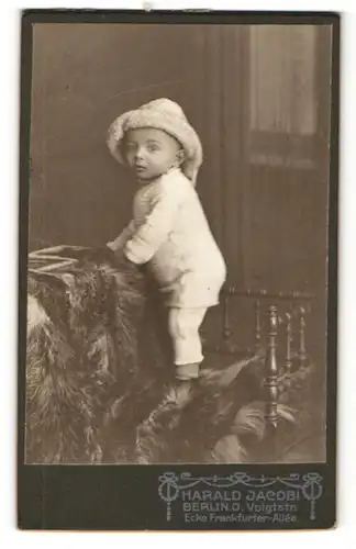 Fotografie Harald Jacobi, Berlin-O, Portrait niedliches Kleinkind in hübscher Kleidung mit Hut auf Fell stehend