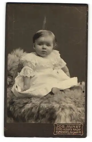 Fotografie Jos. Minet, Elmshorn, Baby im Taufkleid auf Felldecke sitzend