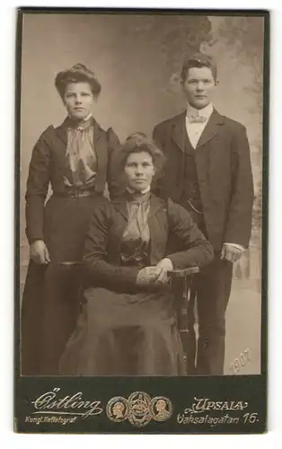 Fotografie Olof Östling, Upsala, Zwei Frauen in Kleidern eine stehend eine sitzend und ein junger Mann im Anzug stehend