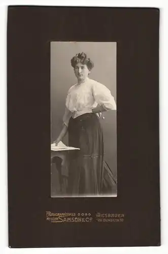 Fotografie Samson & Co., Wiesbaden, Portrait Dame in weisser Bluse mit Dutt u. Zeitung an Tisch gelehnt