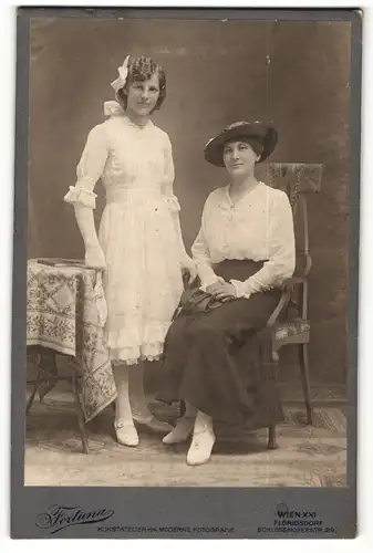 Fotografie Atelier Fortuna, Wien, Portrait Dame in weisser Bluse mit Hut auf Stuhl sitzend u. junge Frau im weissen Kleid