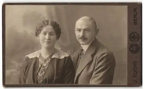 Fotografie J. Fuchs, Berlin, Frau im Kleid mit geflochtenen Haaren mit Mann im Anzug und Halbglatze