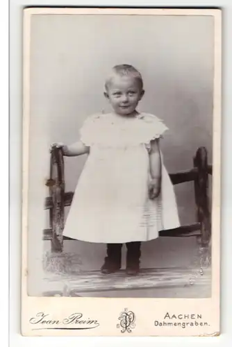 Fotografie Jean Preim, Aachen, Portrait niedliches Kleinkind im weissen Kleid an Zaun gelehnt