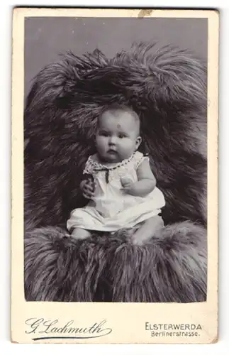 Fotografie G. Lachmuth, Elsterwerda, Portrait niedliches Baby im weissen Kleid auf Fell sitzend