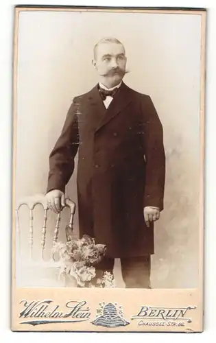 Fotografie Wilhelm Stein, Berlin, Mann im Anzug stehend mit Fliege und breitem Schnauzer