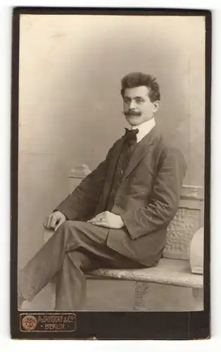 Fotografie A. Jandorf & Co., Berlin, Portrait charmanter Herr im Anzug mit Fliege auf Bank sitzend