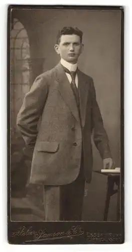 Fotografie Samson & Co., Essen a / Ruhr, Portrait junger Herr im Anzug mit Krawatte an Tisch gelehnt