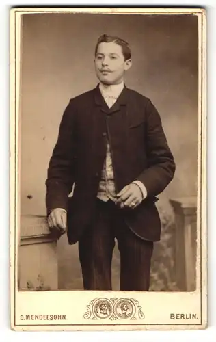 Fotografie D. Mendelsohn, Berlin, Portrait elegant gekleideter Herr mit Zigarre an Sockel gelehnt