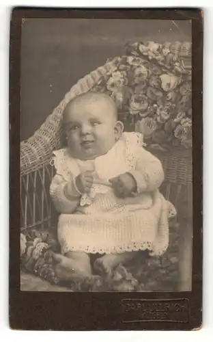 Fotografie Carl Ullrich, Mügeln, Portrait niedliches Baby im hübschen Kleid auf Korbstuhl sitzend