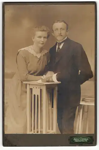 Fotografie Adolf Zierold, Zschopau i / S., Portrait modisch gekleidetes Paar mit Heft an Tisch gelehnt