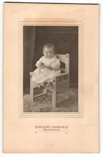 Fotografie Richard Bassler, Oberoderwitz, Portrait sitzendes Kleinkind im weissen Kleid mit Rassel