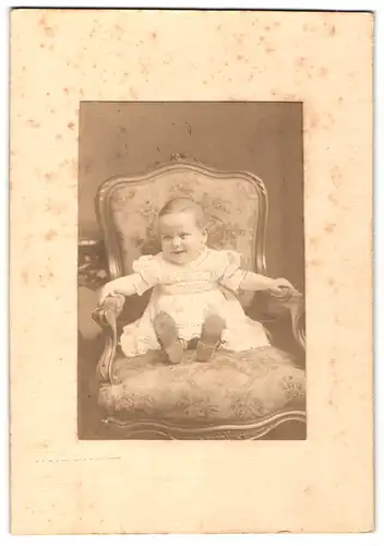 Fotografie unbekannter Fotograf und Ort, Portrait sitzendes Kleinkind im weissen Kleid mit kurzen Haaren