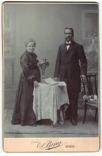 Fotografie Sigmund Bing, Wien, Portrait bürgerlich gekleidetes Paar mit Buch an Tisch gelehnt