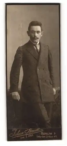 Fotografie Julius Staudt, Berlin, Portrait elegant gekleideter Herr mit Zwicker an Tisch gelehnt