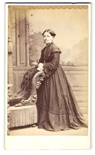 Fotografie W. H. Moore, Bristol, Frau im Kleid stehend mit den Händen auf Sessellehne abgelegt