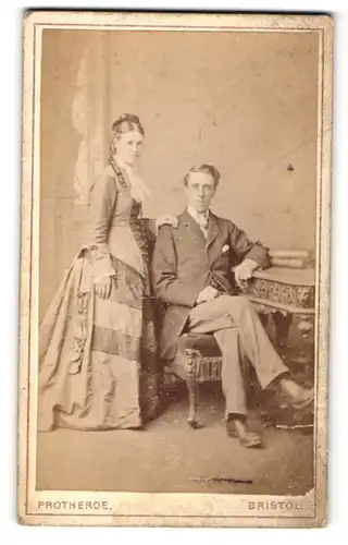 Fotografie Protheroe, Bristol, Mädchen im Kleid stehend neben jungen Mann im Anzug sitzend auf Stuhl