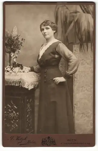 Fotografie J. Krotsch, Wien, Portrait elegant gekleidete Dame mit Blumen an Sockel gelehnt