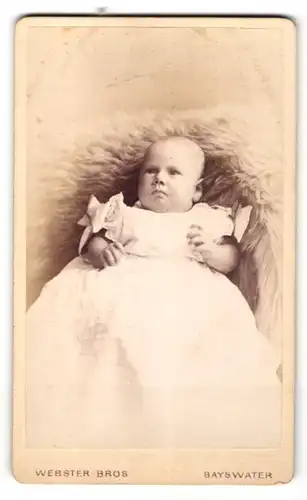 Fotografie Webster Bros., Bayswater, Portrait Baby mit langen Taufkleidchen auf einem Fell liegend