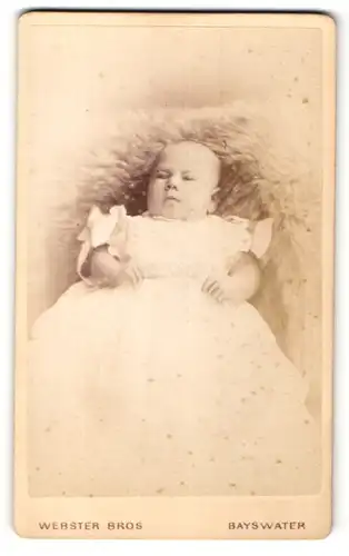 Fotografie Webster Bros., Bayswater, Portrait Baby mit langen Taufkleidchen auf einem Fell liegend