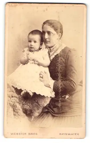 Fotografie Webster Bros., Bayswater, Portrait Baby im weissen Kleidchen im Arm der Mutter
