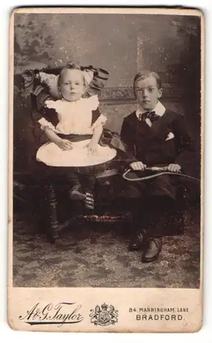 Fotografie A. & G. Taylor, Bradford, Portrait Mädchen im hübschen Kleid und Junge im Anzug mit Peitsche