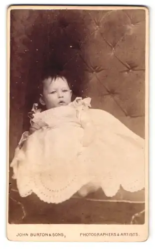 Fotografie John Burton & Sons, Leicester, Portrait niedliches Kleinkind im weissen Kleid auf Sessel sitzend