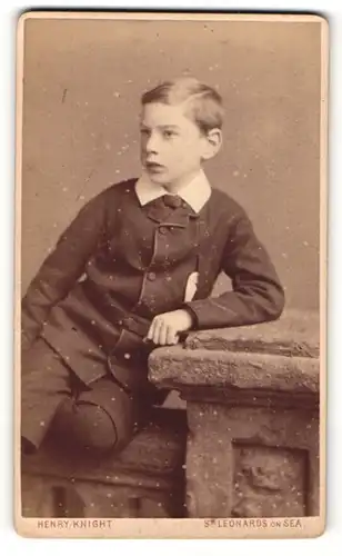 Fotografie Henry Knight, St. Leonards on Sea, Portrait hübsch gekleideter Junge mit Seitenscheitel an Sockel gelehnt