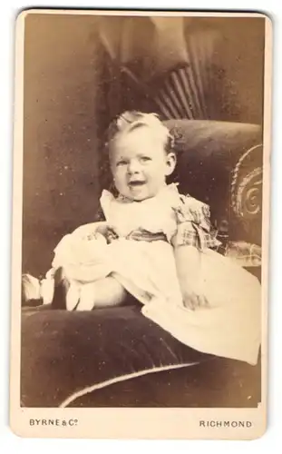 Fotografie Byrne & Co., Richmond, Portrait sitzendes Kleinkind im weissen Kleid mit Schleifchen