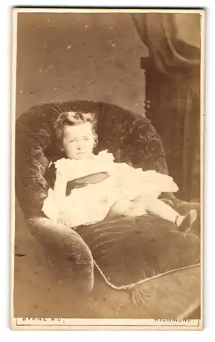 Fotografie Byrne & Co., Richmond, Portrait kleines Mädchen im weissen Kleid auf Sessel sitzend