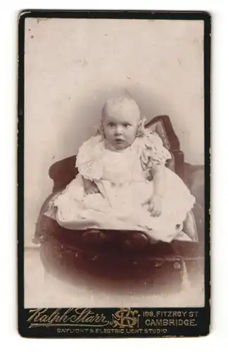 Fotografie Ralph Starr, Cambridge, Portrait sitzendes Baby im hübschen Kleid mit Schühchen