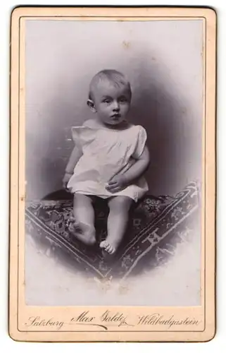 Fotografie Max Balde, Salzburg, Kleinkind in weissem Laibchen sitzt auf einem gewobenem Teppich
