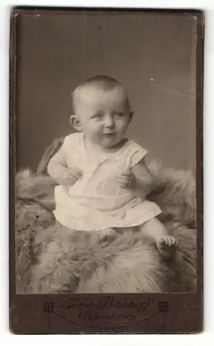 Fotografie Johs. Bischoff, Weinheim, Portrait niedliches Baby im weissen Hemd auf Fell sitzend