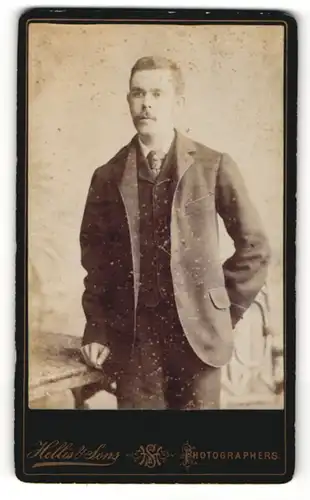 Fotografie Hellis & Sons, London, Portrait bürgerlicher Herr im Anzug mit Krawatte an Tisch gelehnt