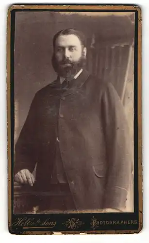 Fotografie Hellis & Sons, London, Portrait bürgerlich gekleideter Herr mit Vollbart an Sockel gelehnt