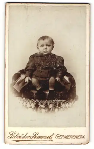 Fotografie Gebr. Rummer, Germersheim, Portrait sitzendes Kleinkind im hübschen Kleid mit kurzen Haaren
