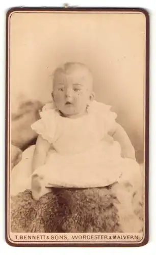 Fotografie T. Bennett & Sons, Worcester, Kleinkind in weissem Kleidchen sitzt mit vorgestrecktem Fuss auf einem Pelz