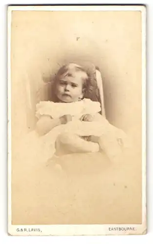 Fotografie G. & R. Lavis, Eastbourne, KLeines Mädchen mit weissem Kleidchen liegt auf gepolstertem Stuhl