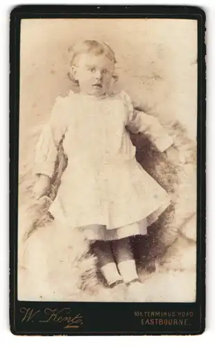Fotografie W. Kent, Eastbourne, Kleines Mädchen in weissem Kleid steht auf mit Fell ausgelegtem Sessel
