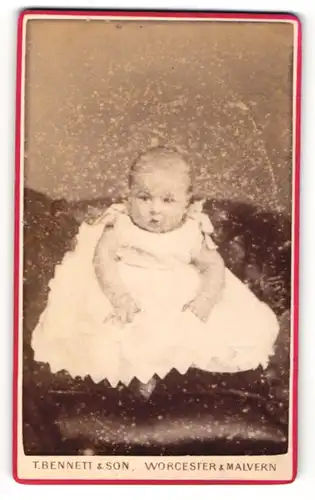 Fotografie T. Bennett & Son, Worcester, Kleinkind in weissem Kleidchen sitzt auf Ledersessel