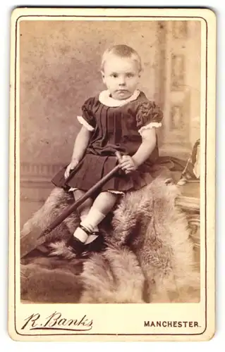 Fotografie R. Banks, Manchester, Portrait sitzendes Mädchen im hübschen Kleid mit Schaufel