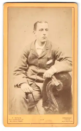 Fotografie R. Banks, Manchester, Portrait sitzender Herr im Anzug mit Einstecktuch und Stock
